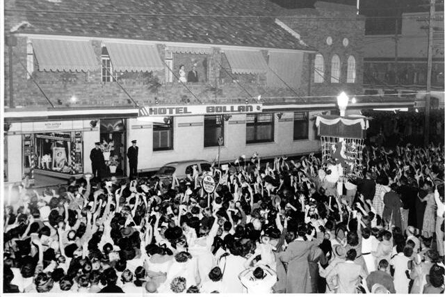 Black and white photo of crowd standing in front of large single story building, two people on a balcony