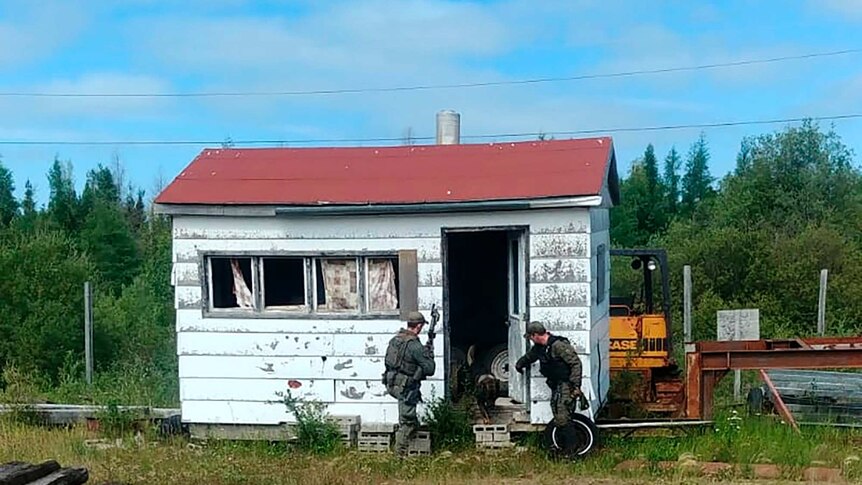 A pixelated image shows two camouflaged police officers entering a dilapidated white wooden shack with a red gable roof.