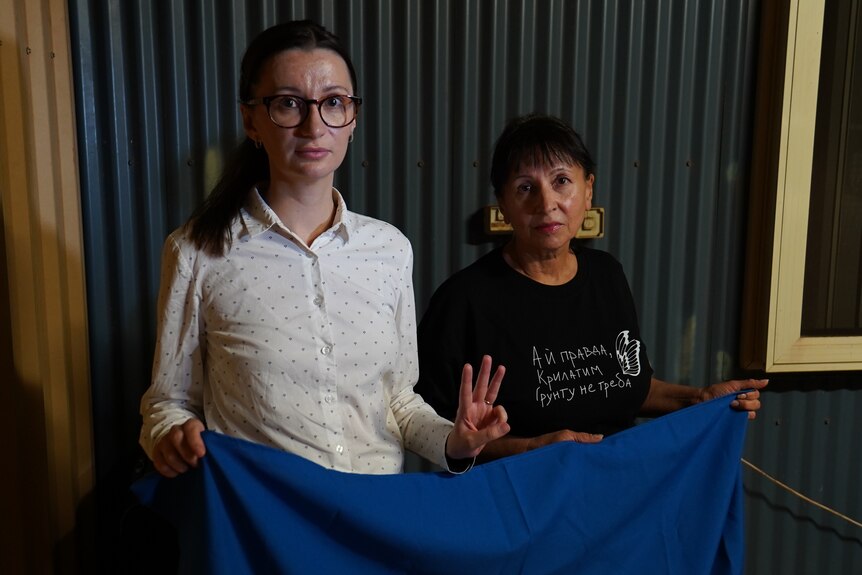Two Ukrainian women hold up a Ukrainian flag at their home. One of the women is holding up three fingers as a peace sign.