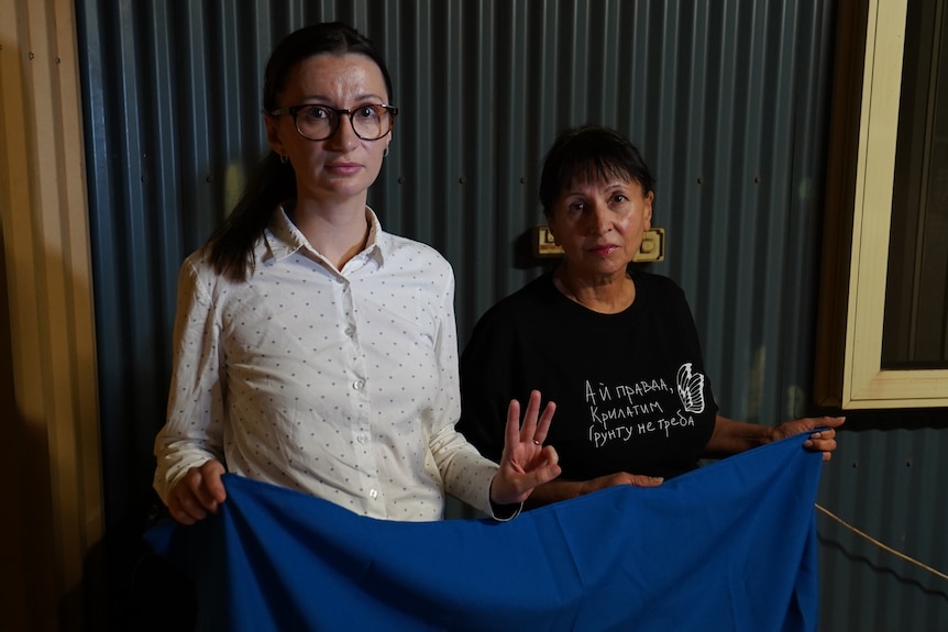Two Ukrainian women hold up a Ukrainian flag at their home. One of the women is holding up three fingers as a peace sign.
