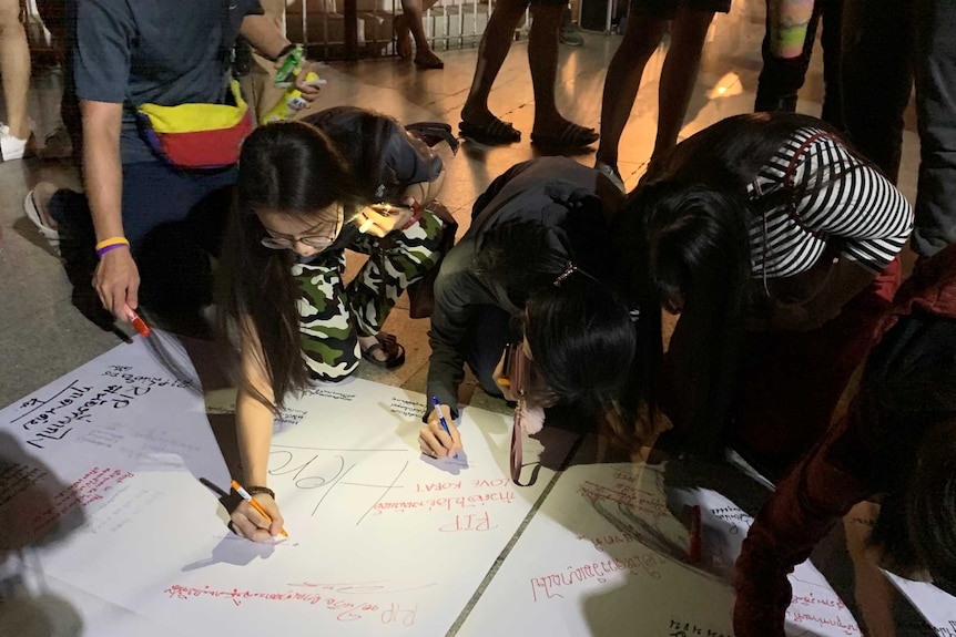 Young Thai people bend down and scrawl messages in texta on large pieces of paper on the ground.