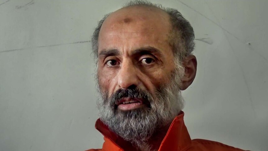 Older man with grey and black beard sits wearing orange prison uniform in front of dull cream wall.