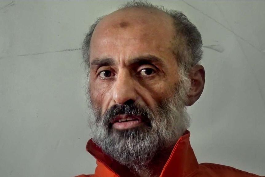 Older man with grey and black beard sits wearing orange prison uniform in front of dull cream wall.