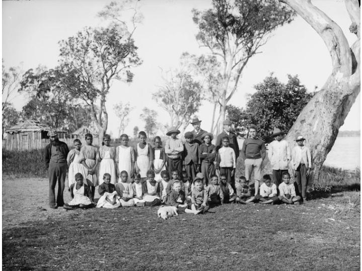 A black and white photo of a group of Aboriginal schoo lchildren dressed in European clothing outside in front of gumtrees