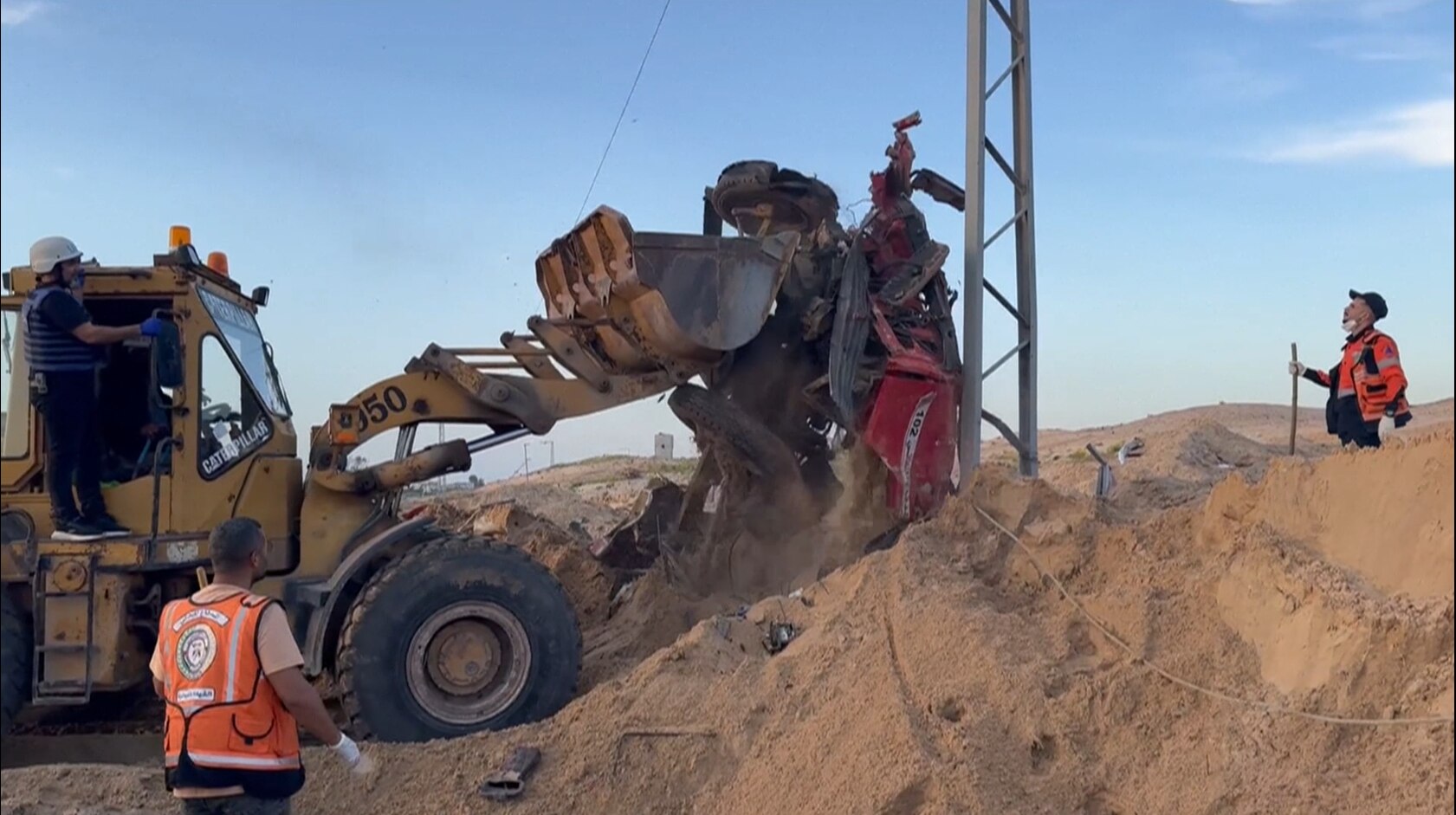 A digger turning over the bombed-out remains of a car beside a road.