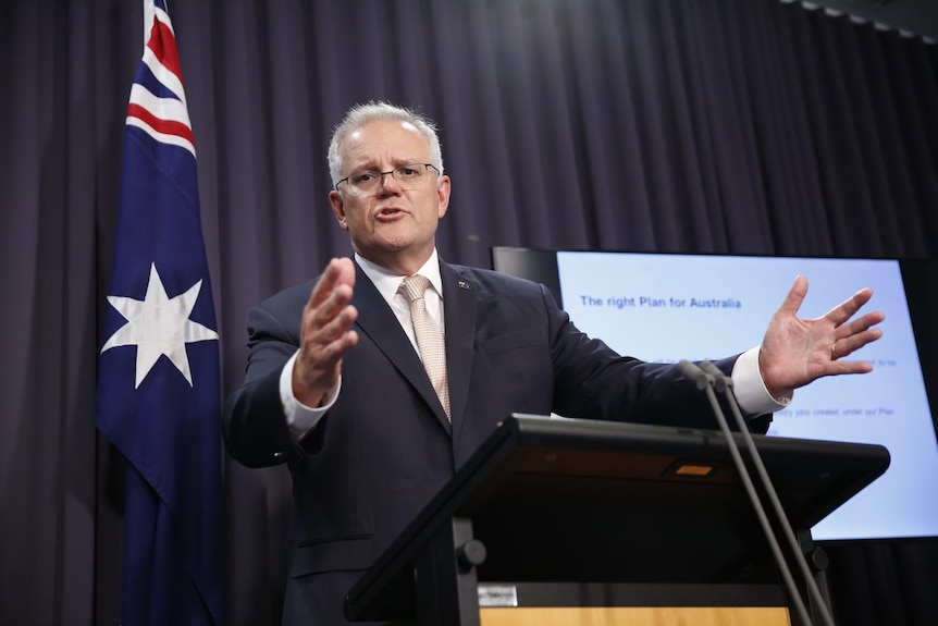 Scott Morrison speaks before a podium with outstretched arms, with a presentation beside him.