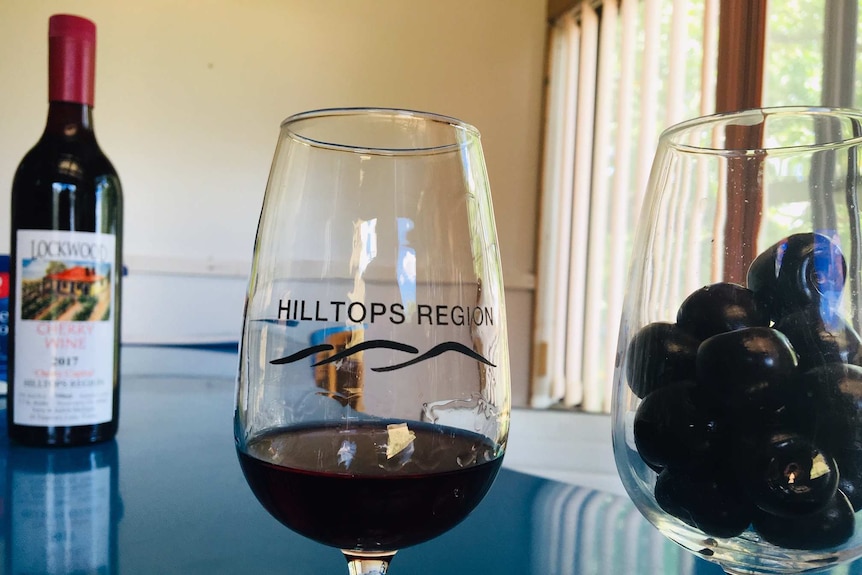 A glass of cherries next to a glass of red cherry wine, with a bottle of cherry wine in the background.