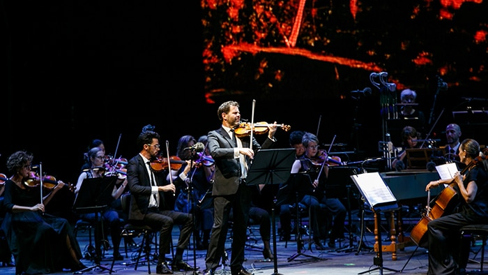 The Sydney Symphony Orchestra performs on stage with a violinist standing up the front and a screen with lava scenes behind them
