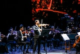 The Sydney Symphony Orchestra performs on stage with a violinist standing up the front and a screen with lava scenes behind them