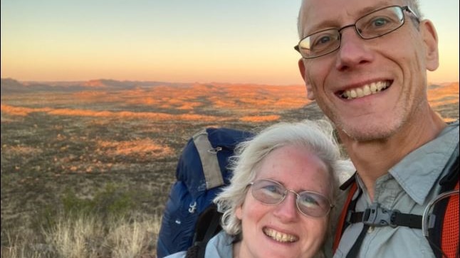 A couple in their 50s smile as they pose for a selfie on a hiking trail with sunlit hills behind them