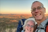 A couple in their 50s smile as they pose for a selfie on a hiking trail with sunlit hills behind them