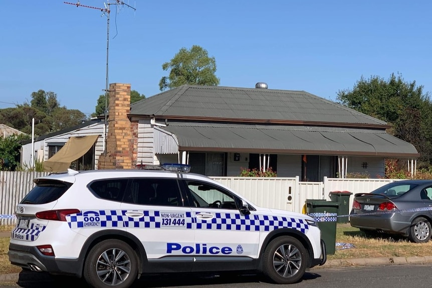 A police vehicle parked in front of a house with a grey tin roof, white weatherboard and white fencing. 