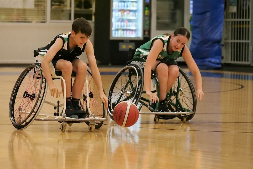 Ellie and a young boy pushing hard in their chairs to reach the basketball.