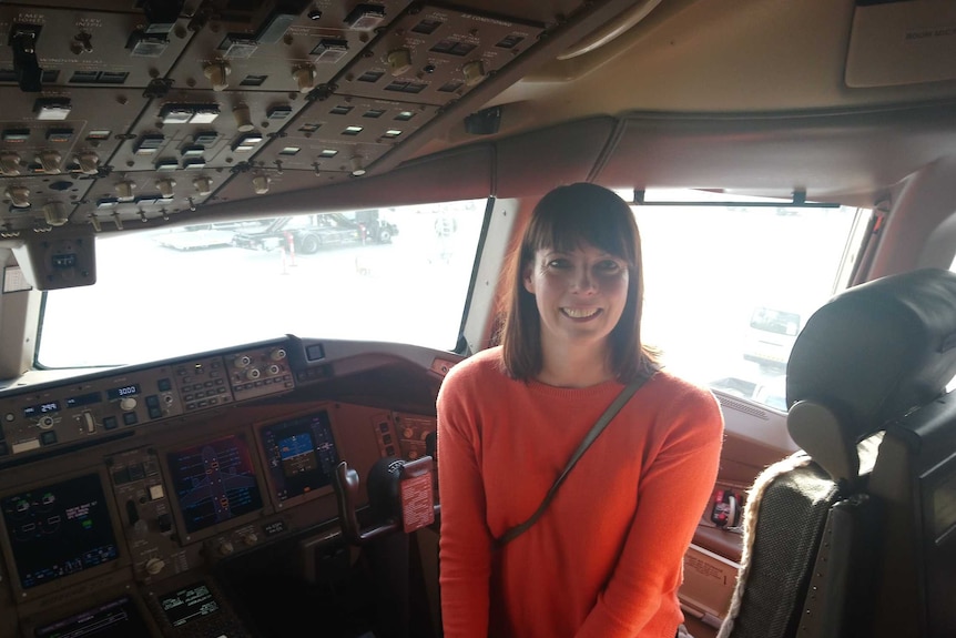 A woman wearing a orange sweater sits in the cockpit of a commercial jet.