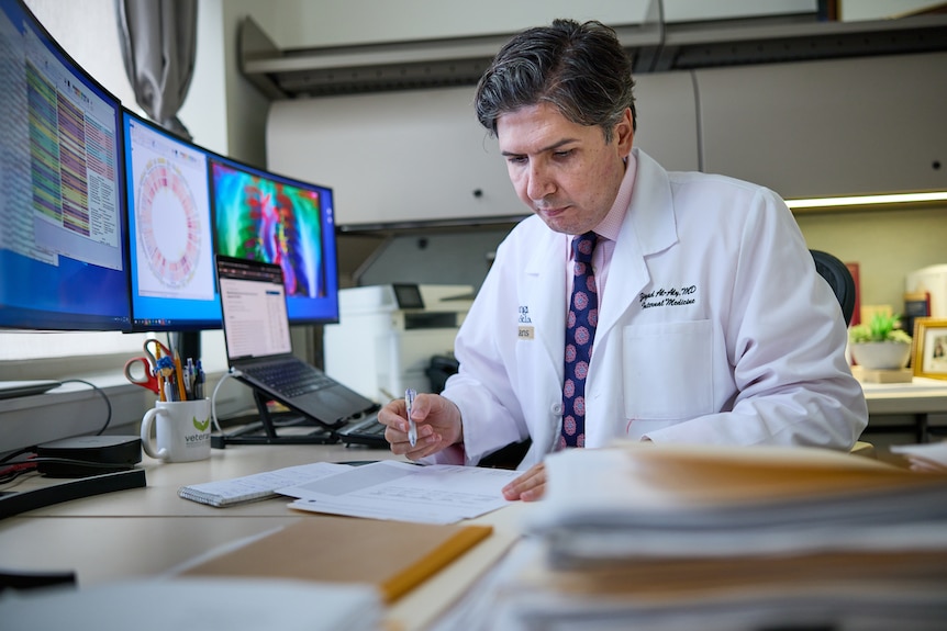 Dr Al-Aly is wearing a white lab coat and reading papers in front of computer screens