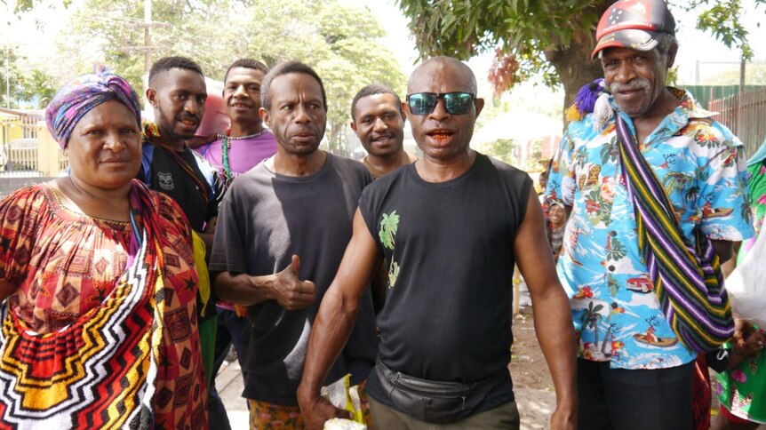 Siedem osób, sześciu mężczyzn i jedna kobieta, stoi przed kamerą na ulicy w Papui-Nowej Gwinei.  Dwóch nosi jaskrawe pilomy. 