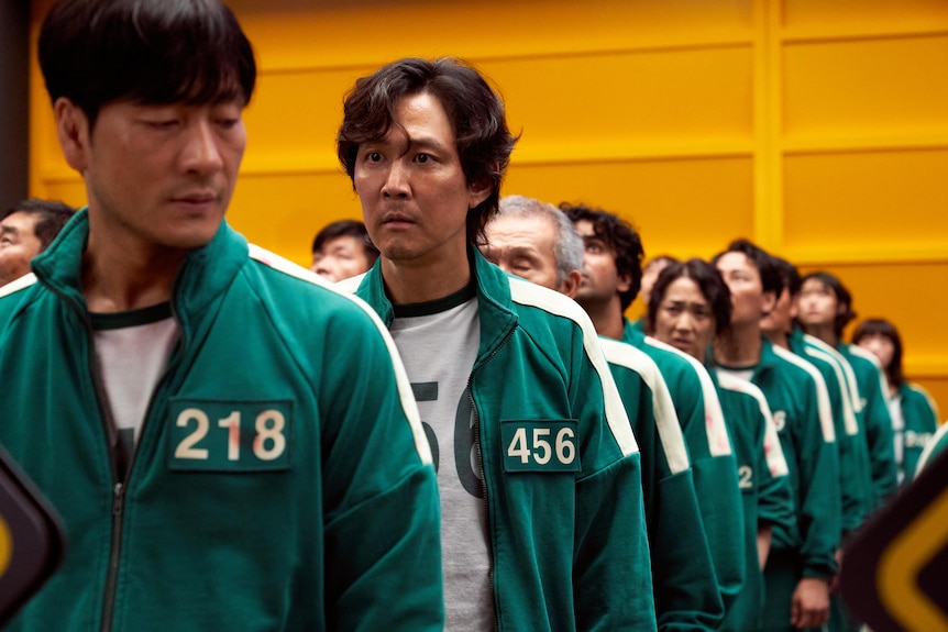 Lee Jung-Jae stands in line in a green tracksuit with a number on it, looking worried