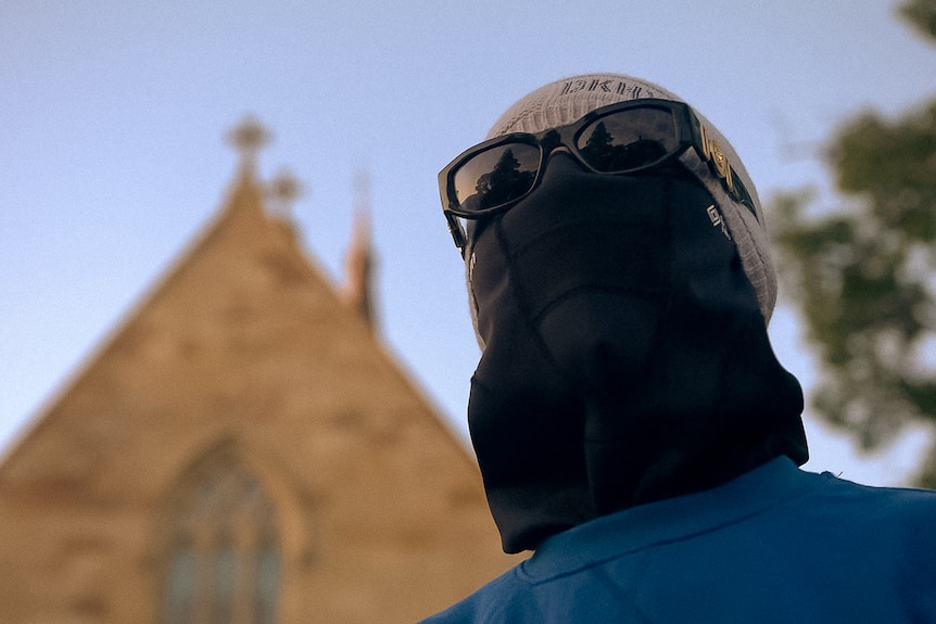 A woman wearing a beanie, mask and glasses, stands in front of a sandstone church building.