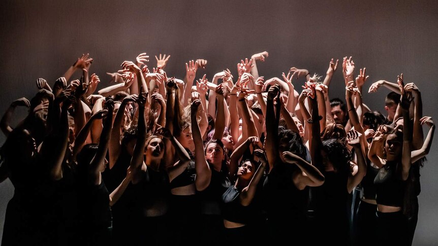 On stage a large group of dancers in black raise their bare arms, reaching up