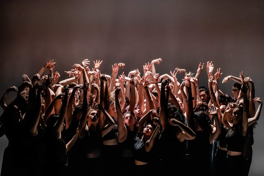 On stage a large group of dancers in black raise their bare arms, reaching up