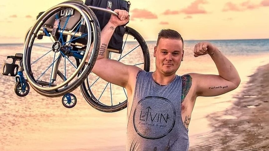 A young man standing on a beach flexes his arms while holding up a wheelchair.