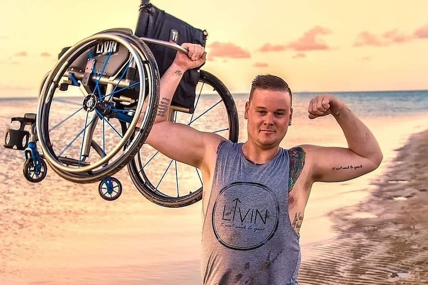 A young man standing on a beach flexes his arms while holding up a wheelchair.