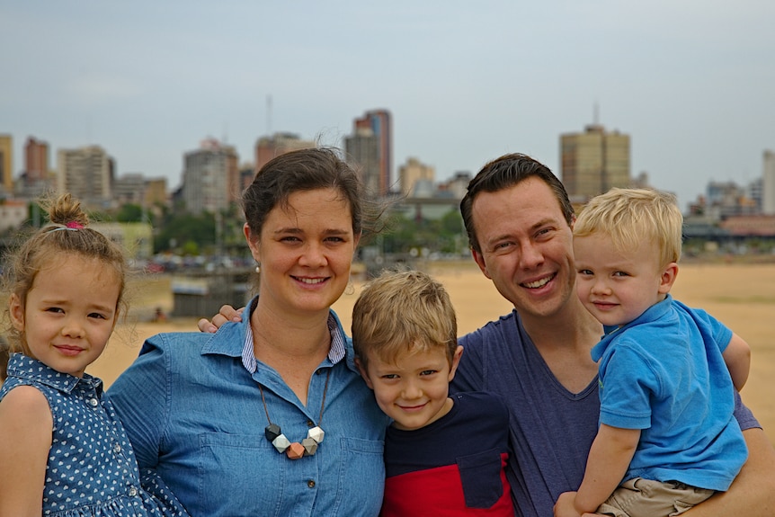 Two middle-aged parents hold three small fair children in their arms in city park on sunny day.