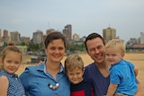 Two middle-aged parents hold three small fair children in their arms in city park on sunny day.