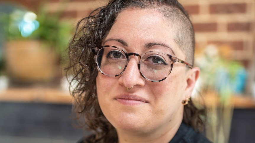 Isabelle Oderberg, with glasses, brown curly hair and half-shaved head, and black t-shirt, smiles with mouth closed.