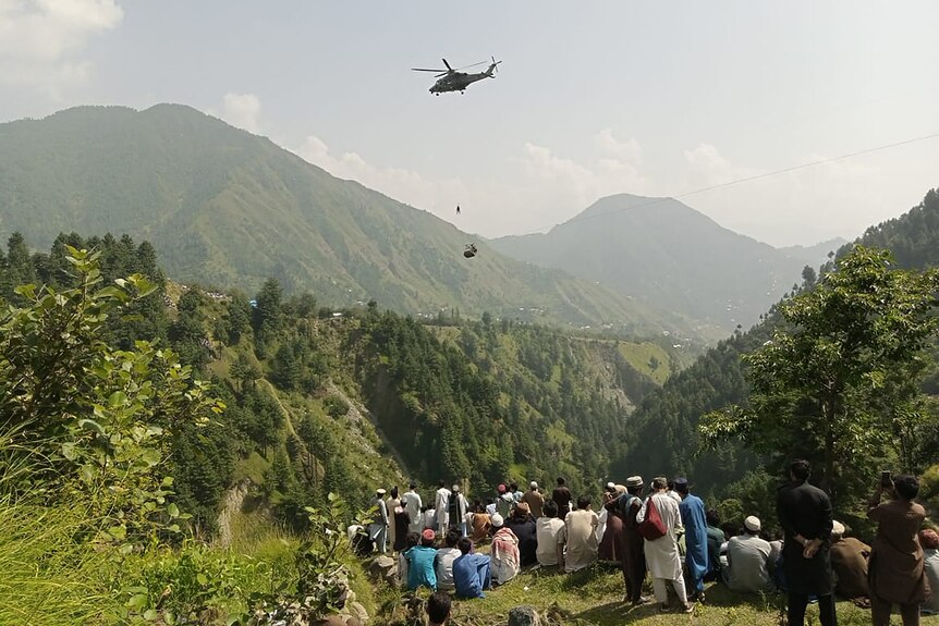 People watch as an army soldier slings down from a helicopter among mountains. 