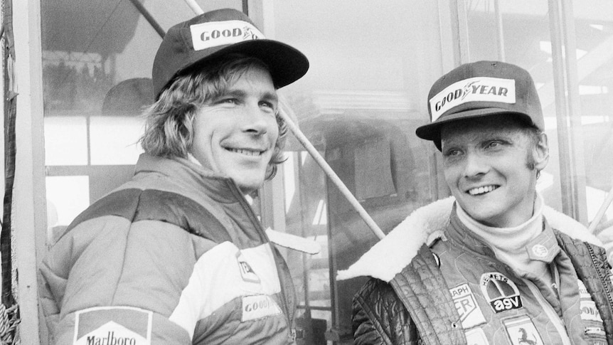 Two Formula One drivers wearing jackets and caps smile as they wait ahead of a race.