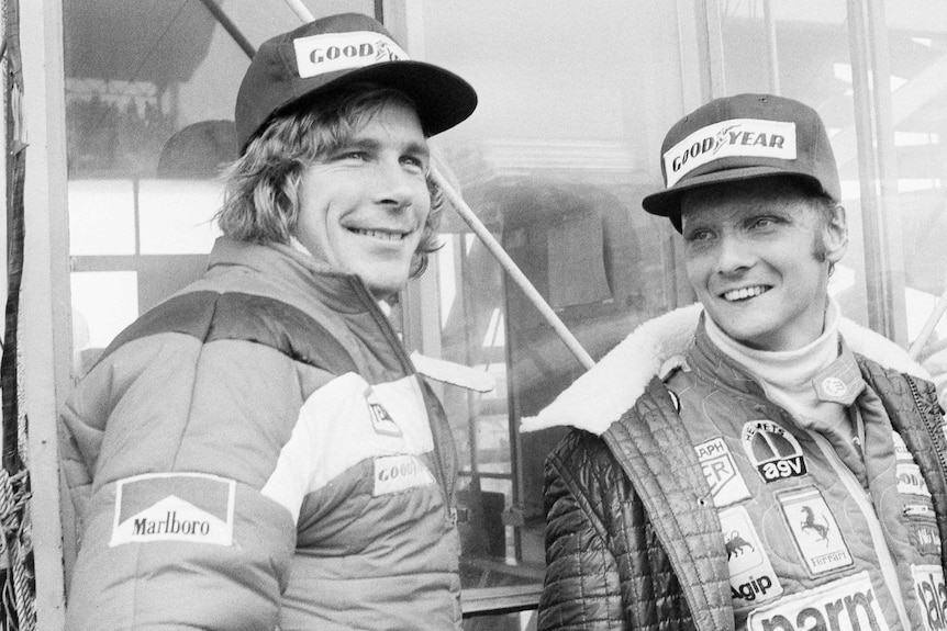 Two Formula One drivers wearing jackets and caps smile as they wait ahead of a race.