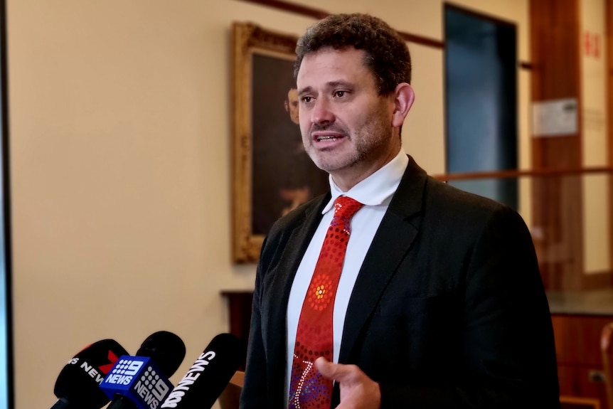 A man with a beard wearing a black suit and red tie speaks to microphones