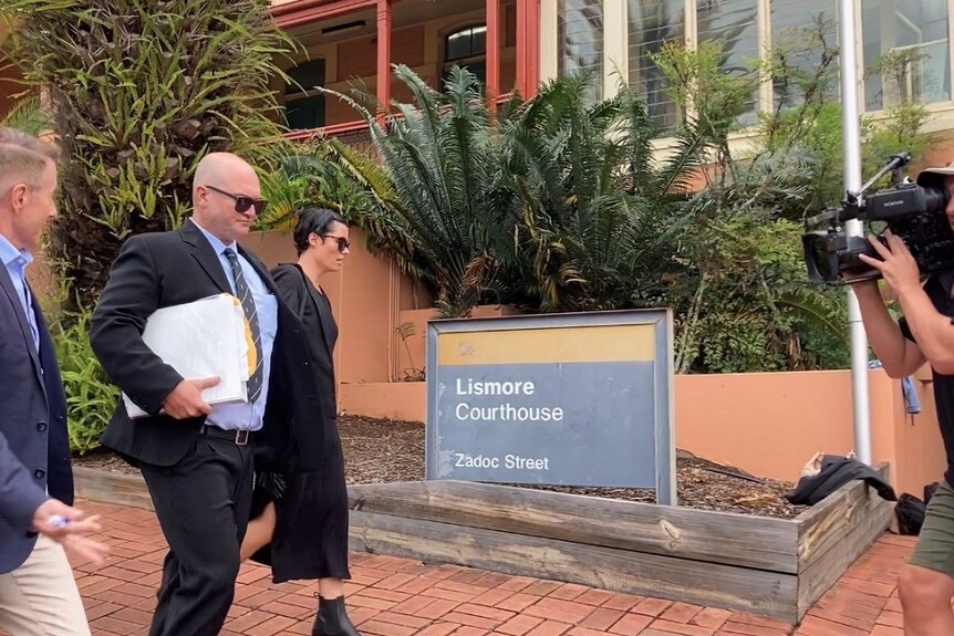 A man in sunglasses and a suit with documents walking next to a woman in front of a court house with media.