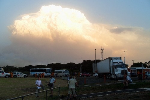 News crew standing on edge of oval with fire trucks in background and huge smoke cloud with orange tinge.