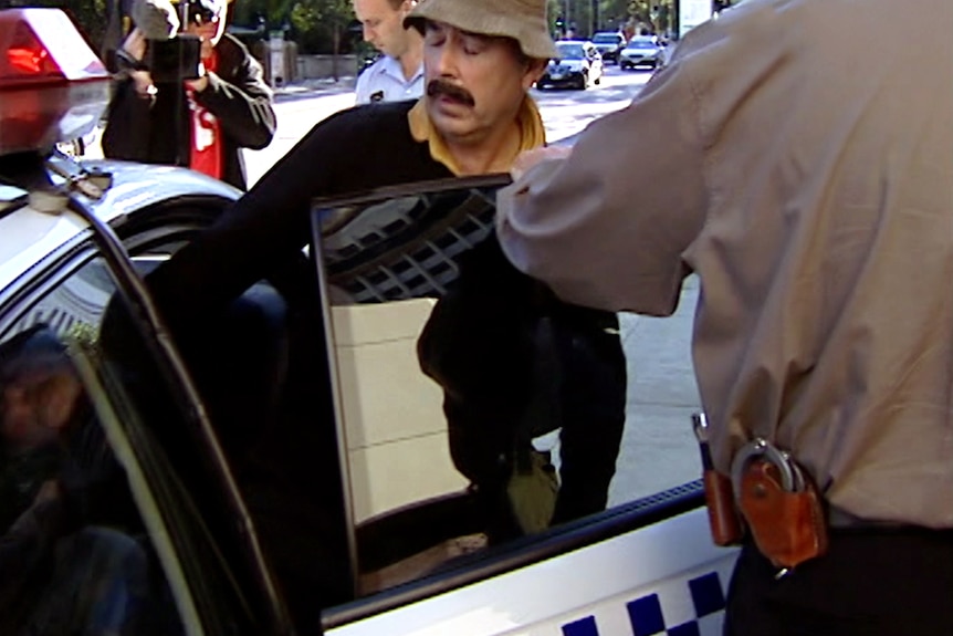 A man with a moustache wearing a bucket hat stepping into the back of a police car.