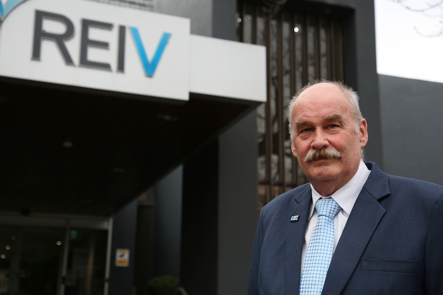 A man with a moustache wearing a light blue tie and navy suit.