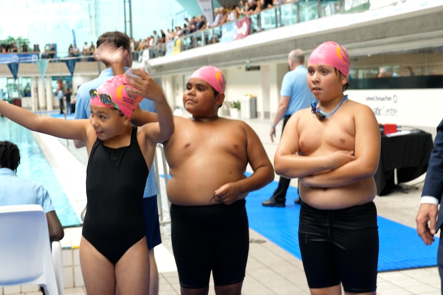 Three kids in swimming costumes wave to the crowd above a pool.