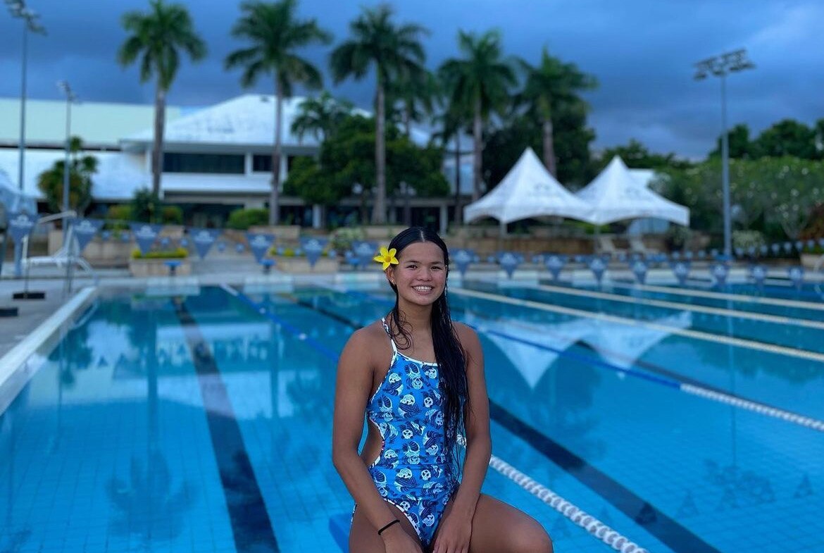 A young girl in a blue swimsuit sits on the edge of a swimming pool with a yellow flower in her hair and palm trees 