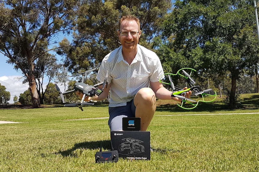 Teacher kneels on grass holding two drones behind three trees.