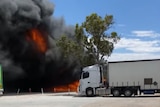 A white semi-trailer with flames and smoking coming from another truck behind it