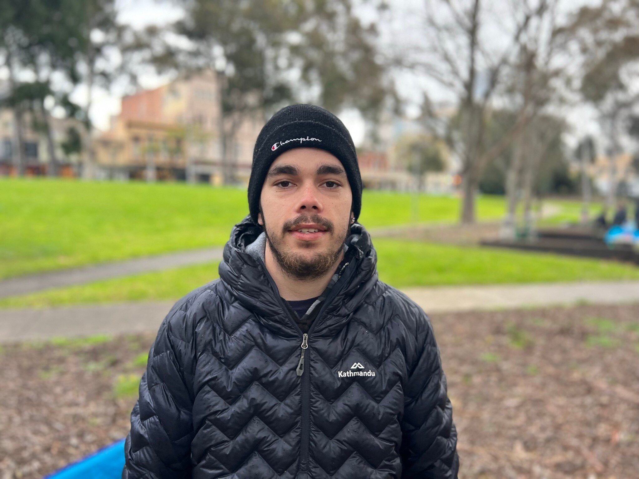 BJ Braybon smiles, standing in a park on a rainy day, dressed in a black beanie and puffer jacket.