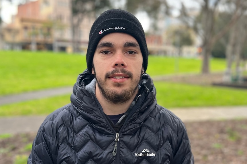 BJ Braybon smiles, standing in a park on a rainy day, dressed in a black beanie and puffer jacket.