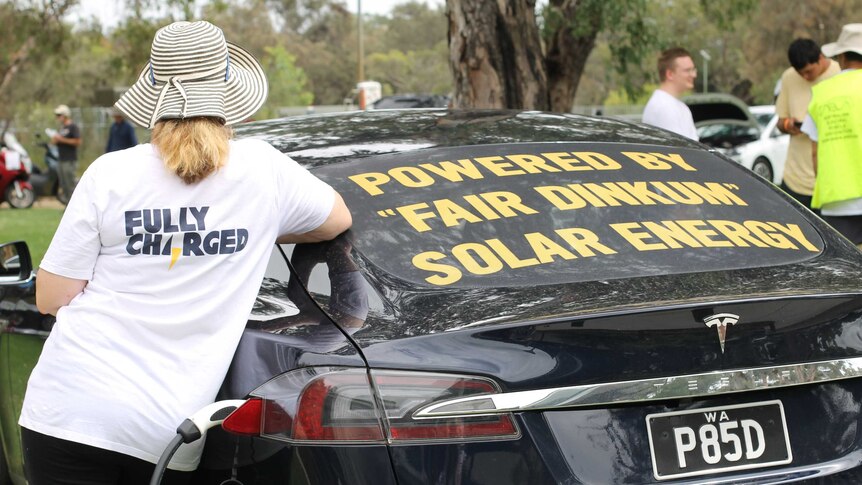 Woman with t shirt on saying "fully charged" leaning on the back of car with sign that says "powered by fair dinkum solar energy