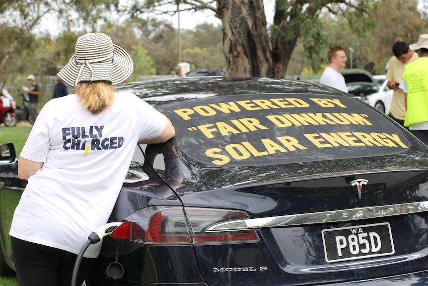 Woman with t shirt on saying "fully charged" leaning on the back of car with sign that says "powered by fair dinkum solar energy