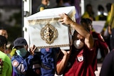 Two people wearing masks carry a white coffin among a crowd of people.