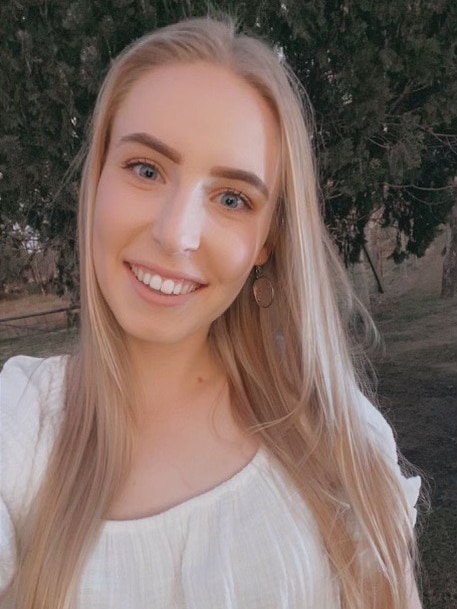 A young woman with blonde hair smiles at the camera, trees behind her