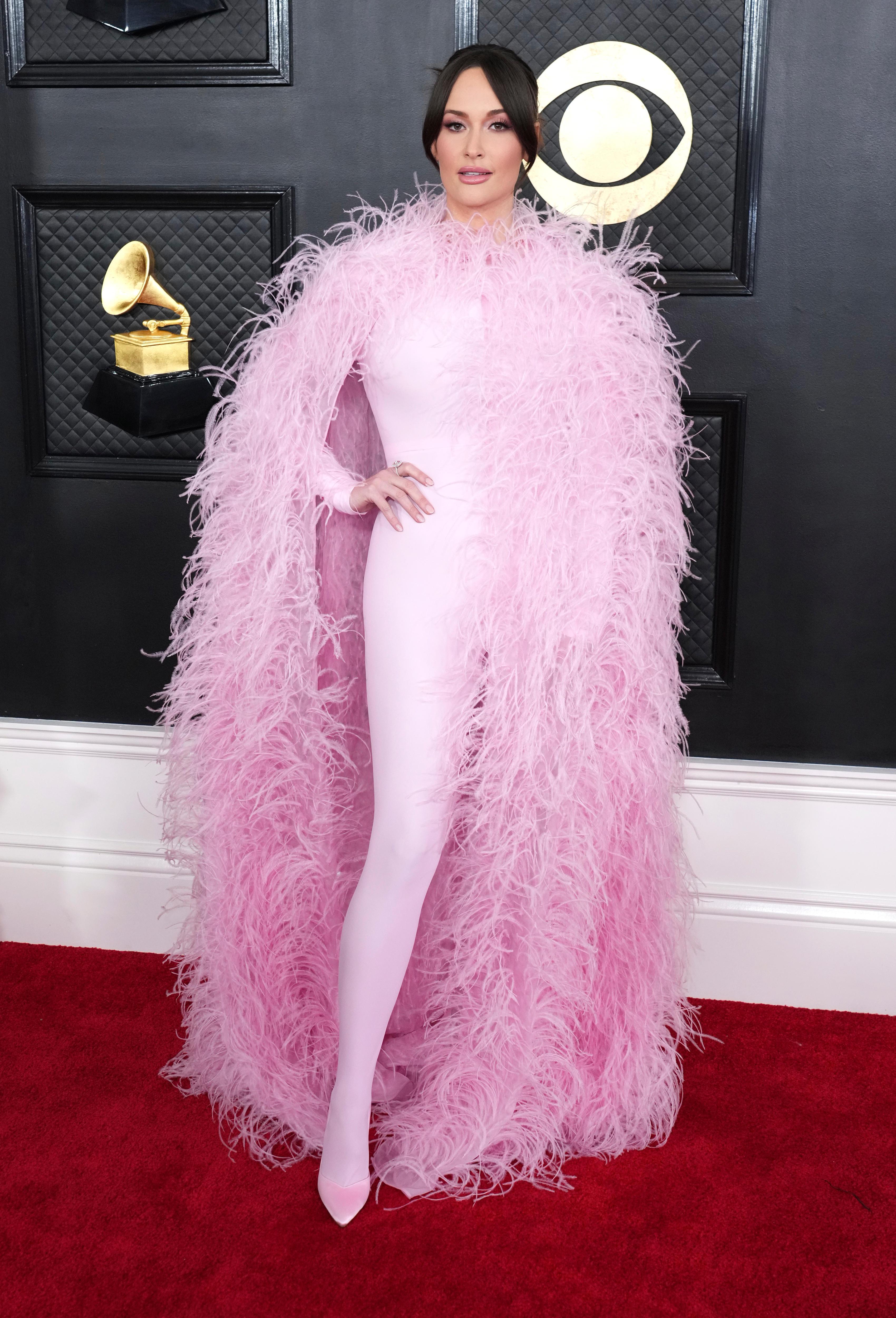 Kacey Musgraves wearing a pale pink fully body leotard with a long fluffy feathered coat. 