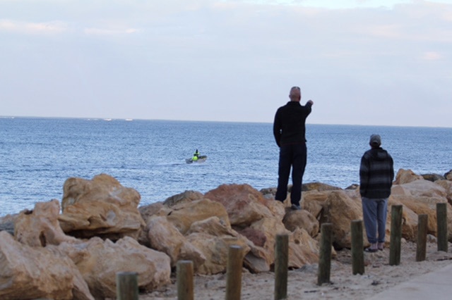 Two men standing near rocks onshore look out to sea, one of them pointing to the horizon as a small boat moves across the water.