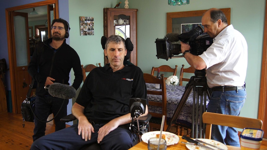 Cameraman and sound recordist holding equipment filming Yerbury in wheelchair.